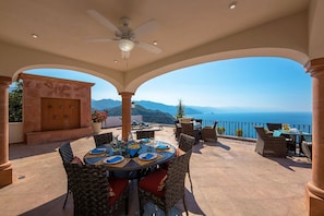 An outdoor patio with a table set for dining, surrounded by wicker chairs, and overlooking a scenic view of the sea and mountains.