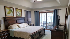 Quarto com uma cama grande, cômoda de madeira, escrivaninha com computador e televisão. Há duas pinturas na parede e uma janela com cortinas.
