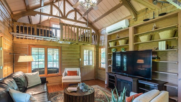 A spacious living room with a large TV, comfortable seating, and a high ceiling with wooden beams.