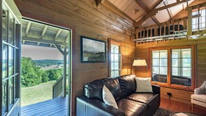 A cozy cabin interior with a leather sofa, a small table, and a view of a green landscape through large windows.