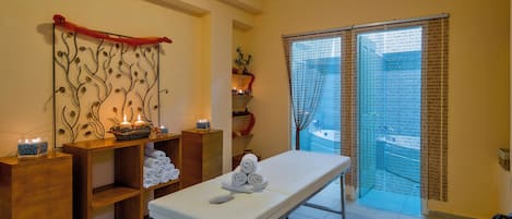 A spa room with a massage table, shelves with towels, decorative wall art, and a view through a window with a curtain.