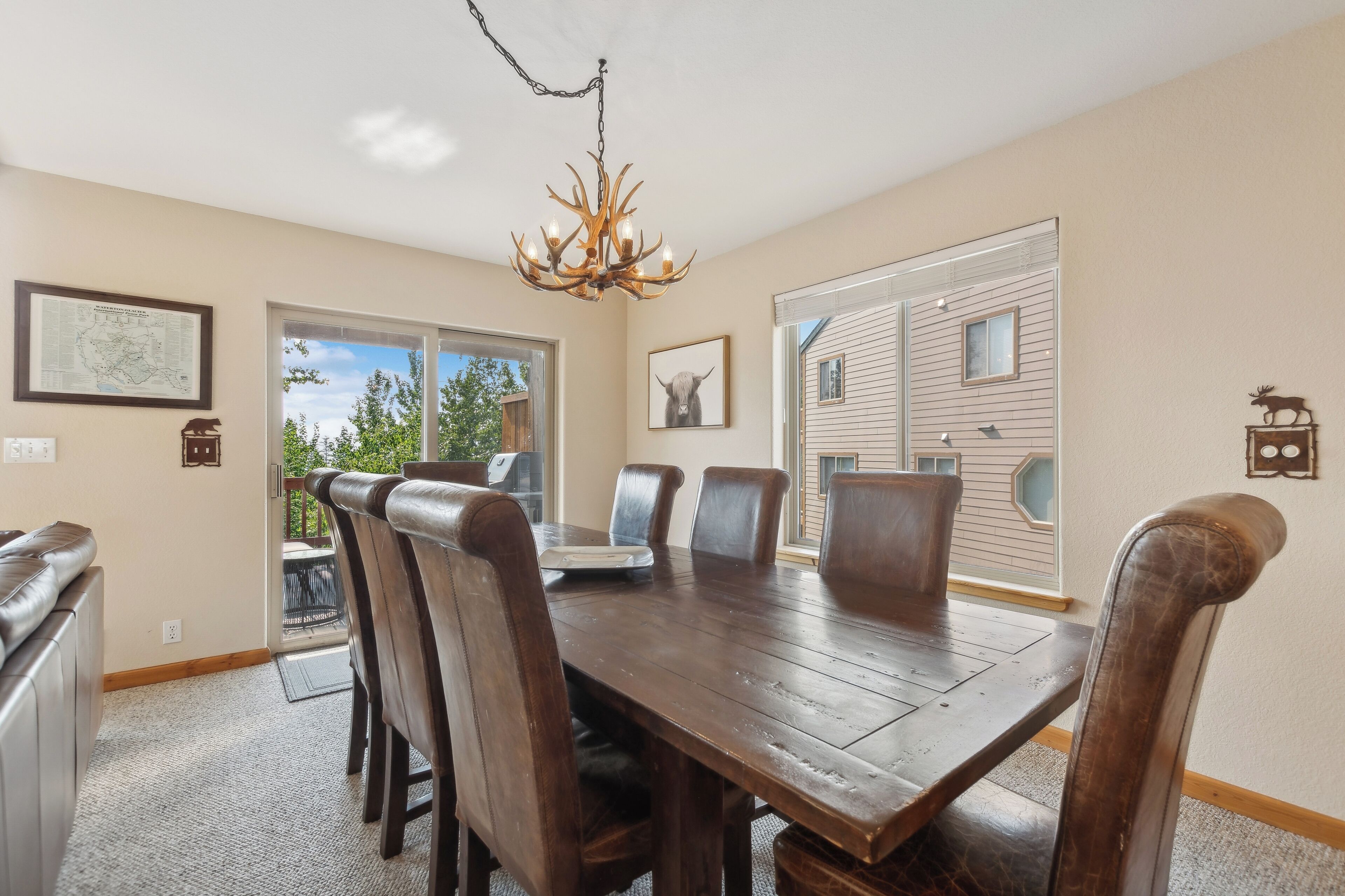 A dining room with a wooden table, leather chairs, a chandelier, and a large window with a view of a house.