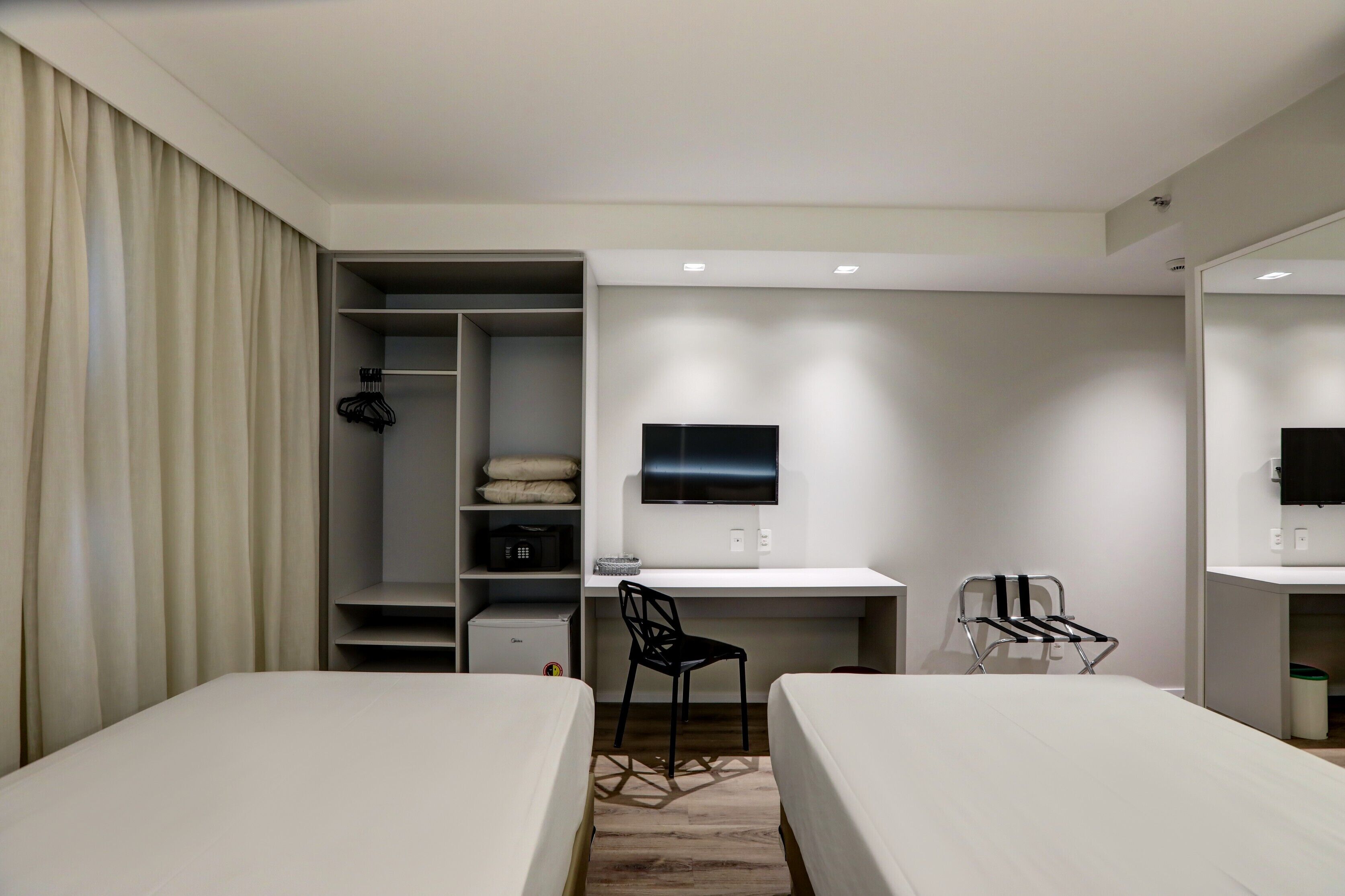 A hotel room with two beds, a desk with a chair, a TV, a wardrobe, and a small kitchenette.