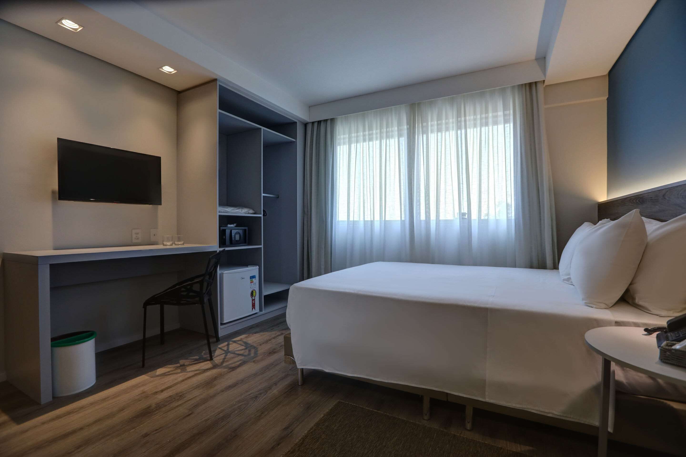 Une chambre d’hôtel avec un lit, un bureau, une chaise, une télévision et une fenêtre avec des rideaux.