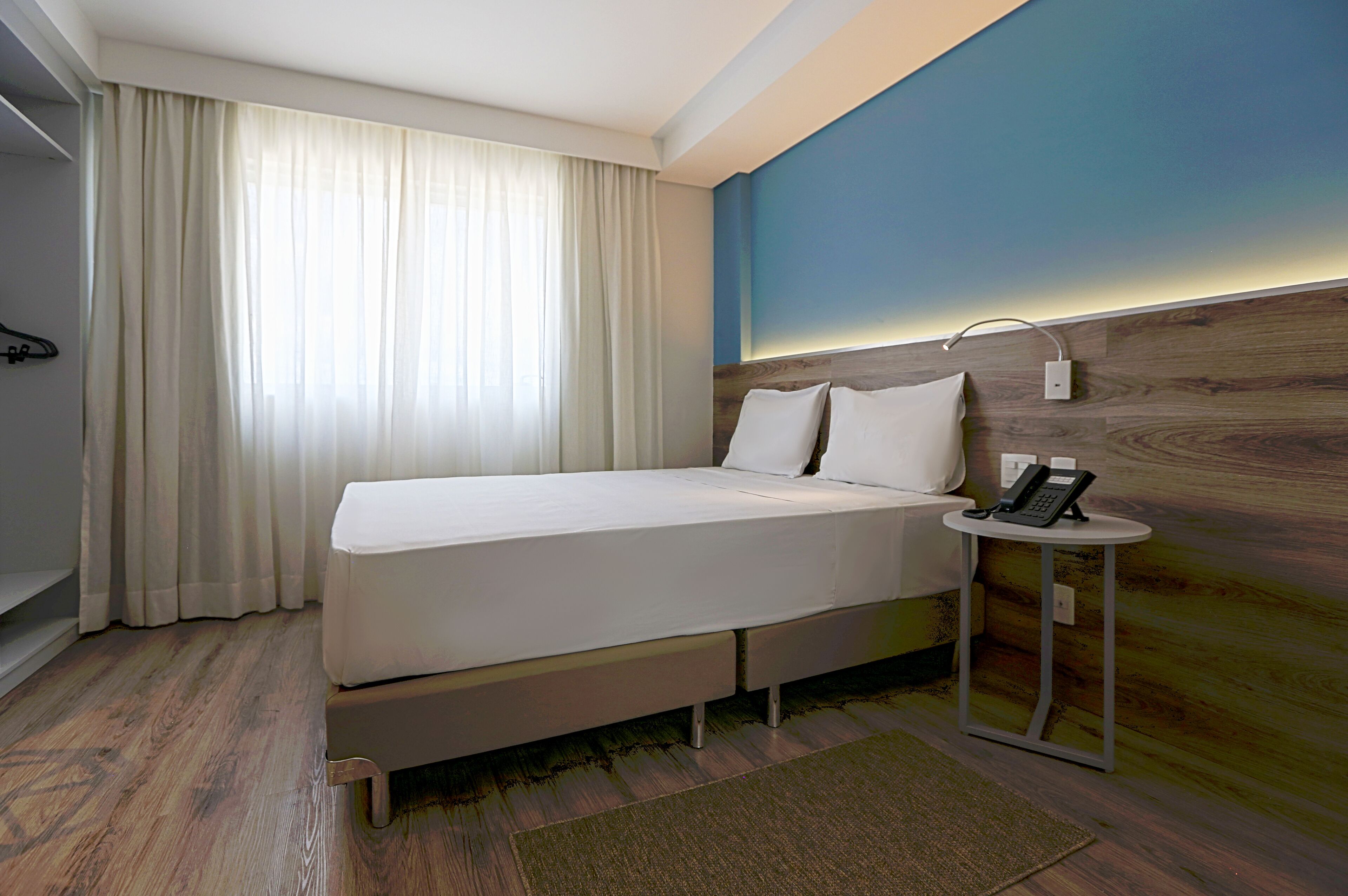 A hotel room with a bed, a bedside table with a phone, a window with curtains, and a wall with a wooden panel.