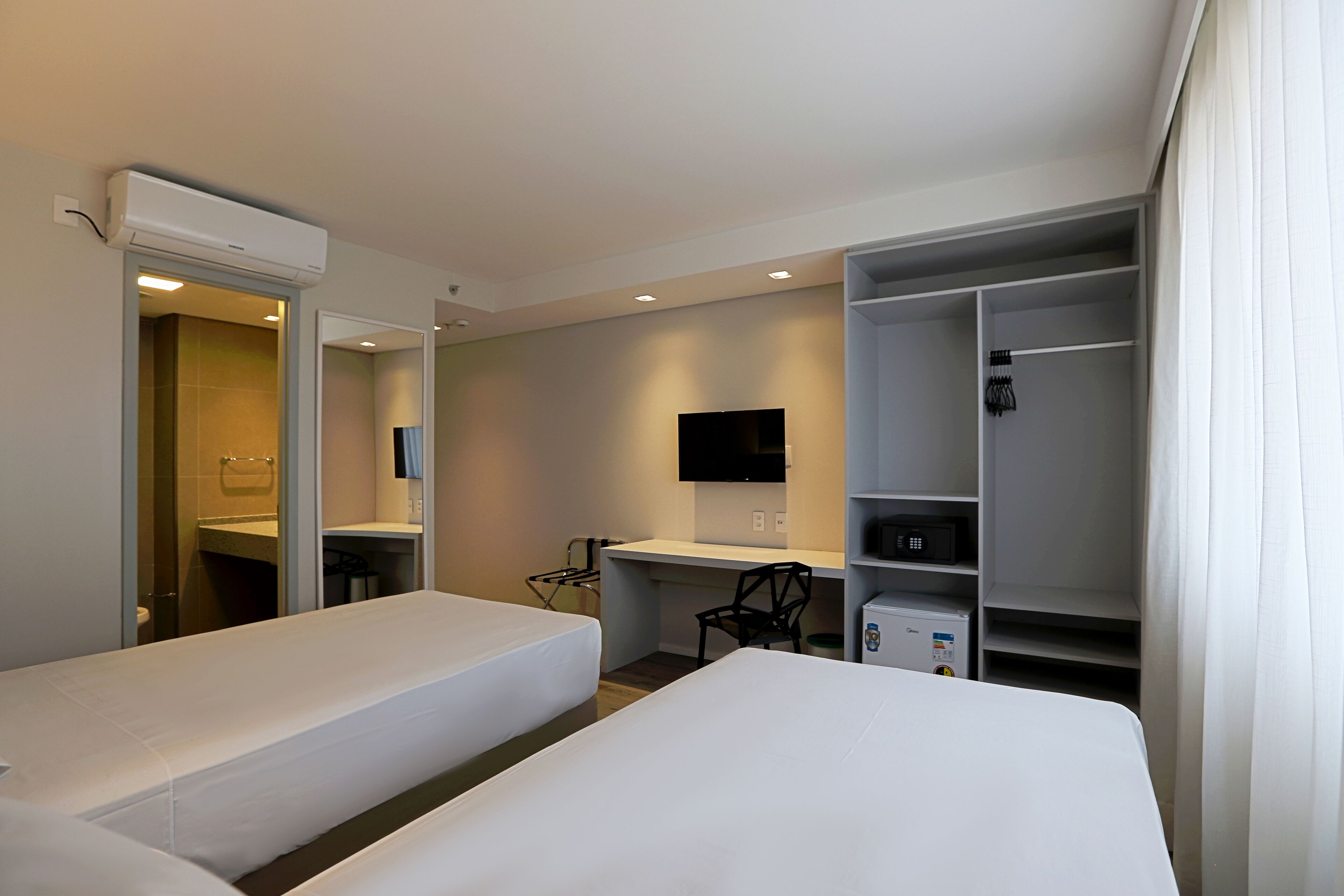 A hotel room with two single beds, a desk with a chair, a TV mounted on the wall, and a wardrobe with shelves.