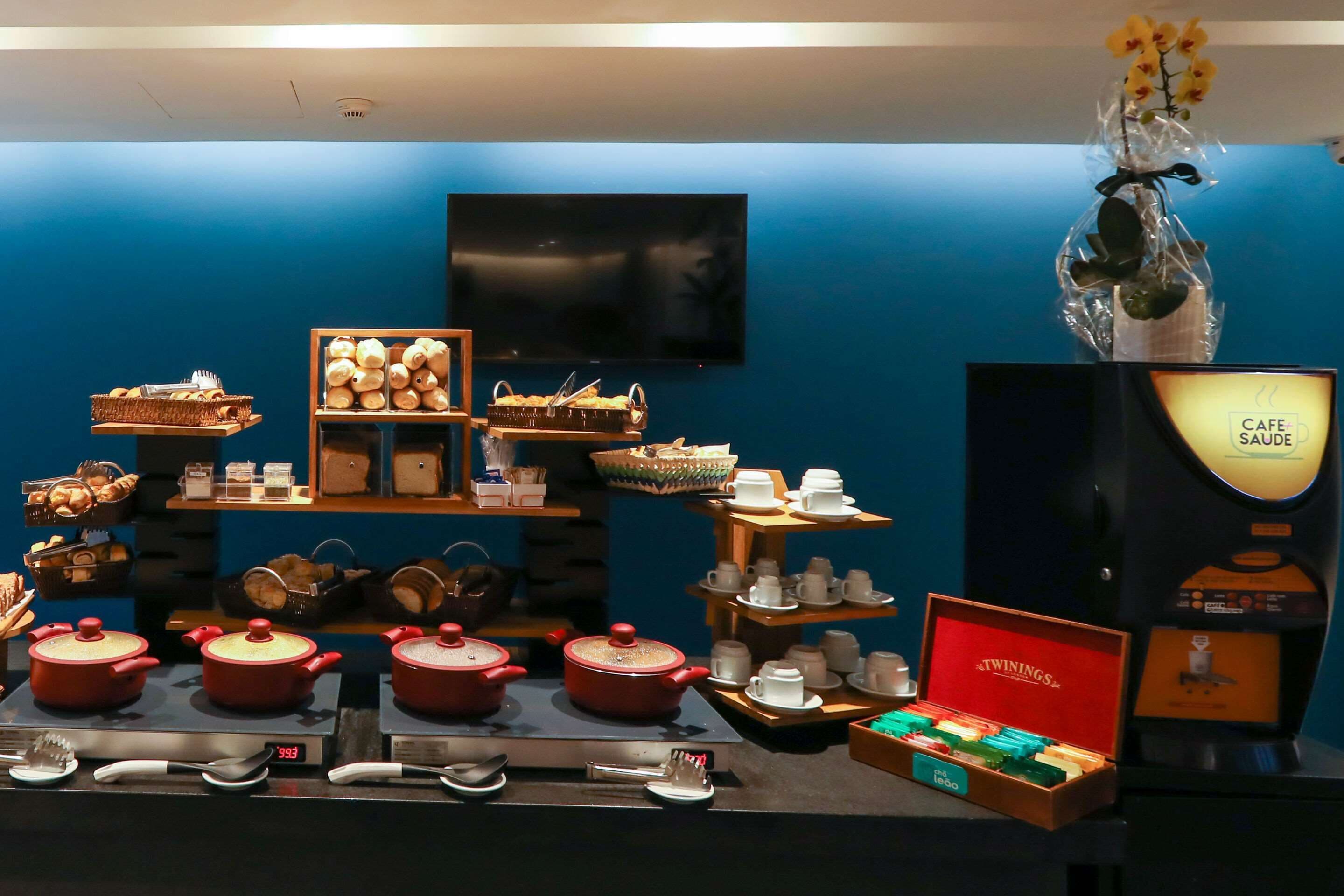 Exposição de utensílios de cozinha e artigos para refeições, incluindo panelas, frigideiras e uma máquina de café, em frente a uma parede azul com uma televisão acima.