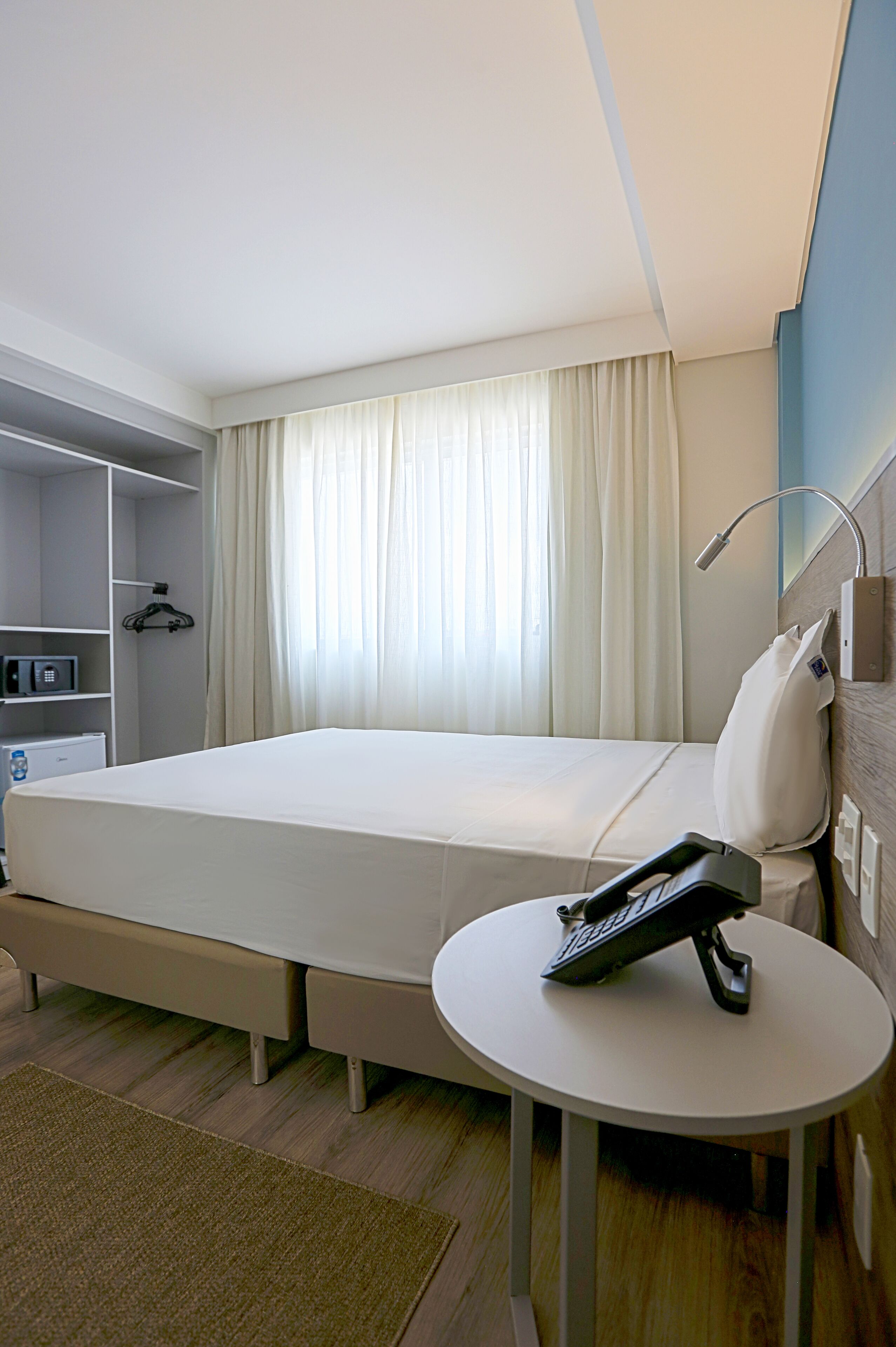 Une chambre d’hôtel avec un lit simple, une table de chevet avec un téléphone, un petit bureau, un four à micro-ondes et une armoire.