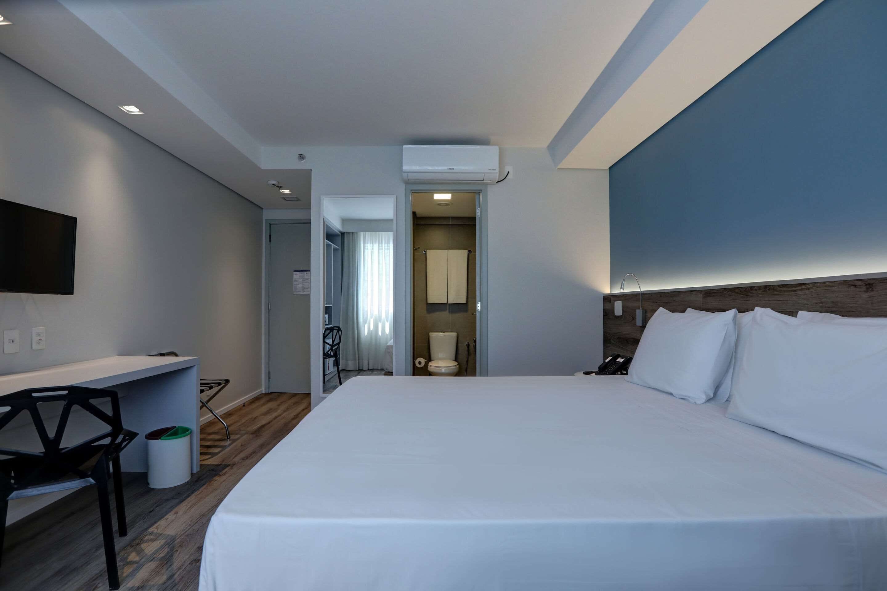 A hotel room with a large bed, a desk, a chair, a TV, and a bathroom visible through an open door.