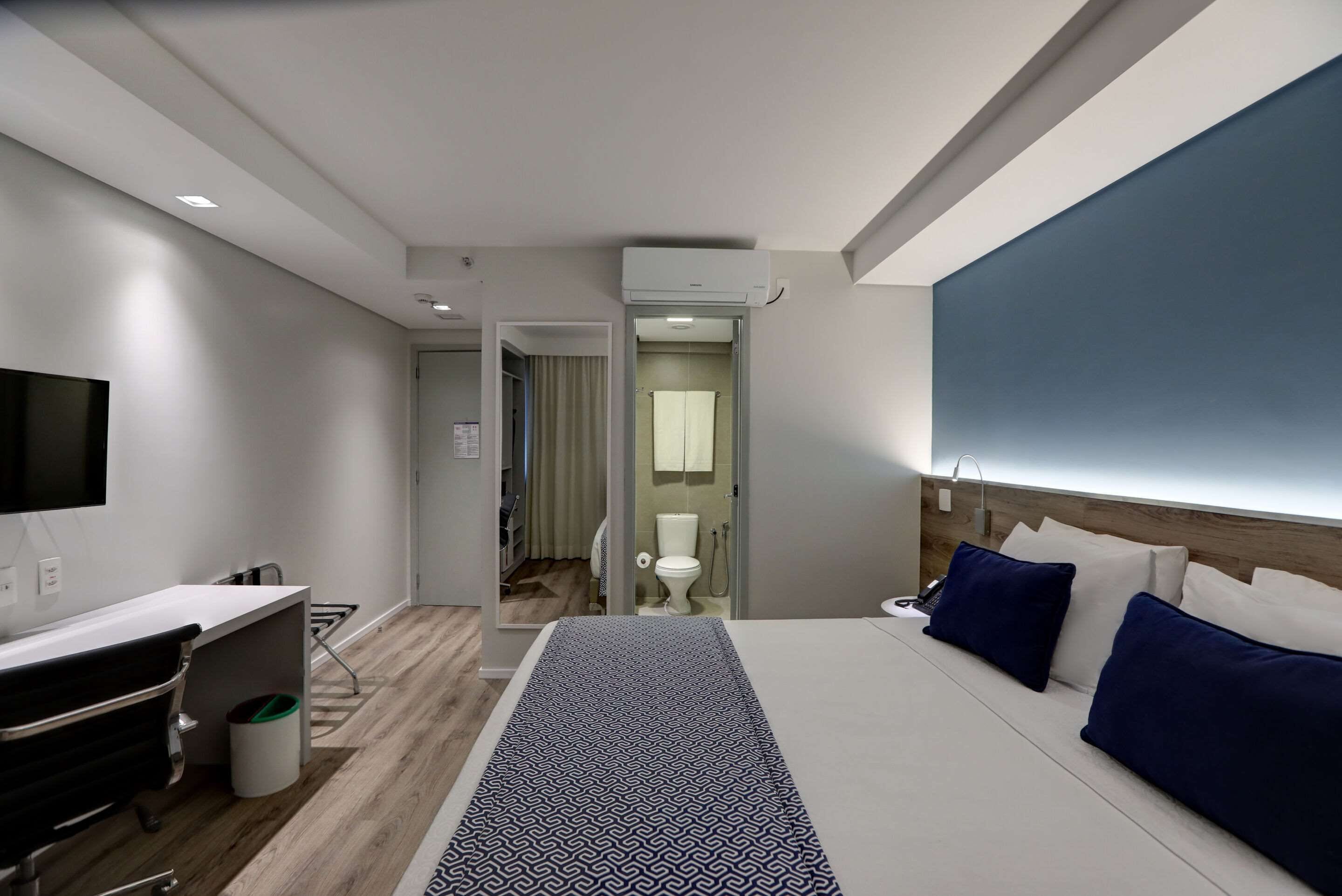 A modern hotel room with a bed, bedside table, and a desk with a chair.