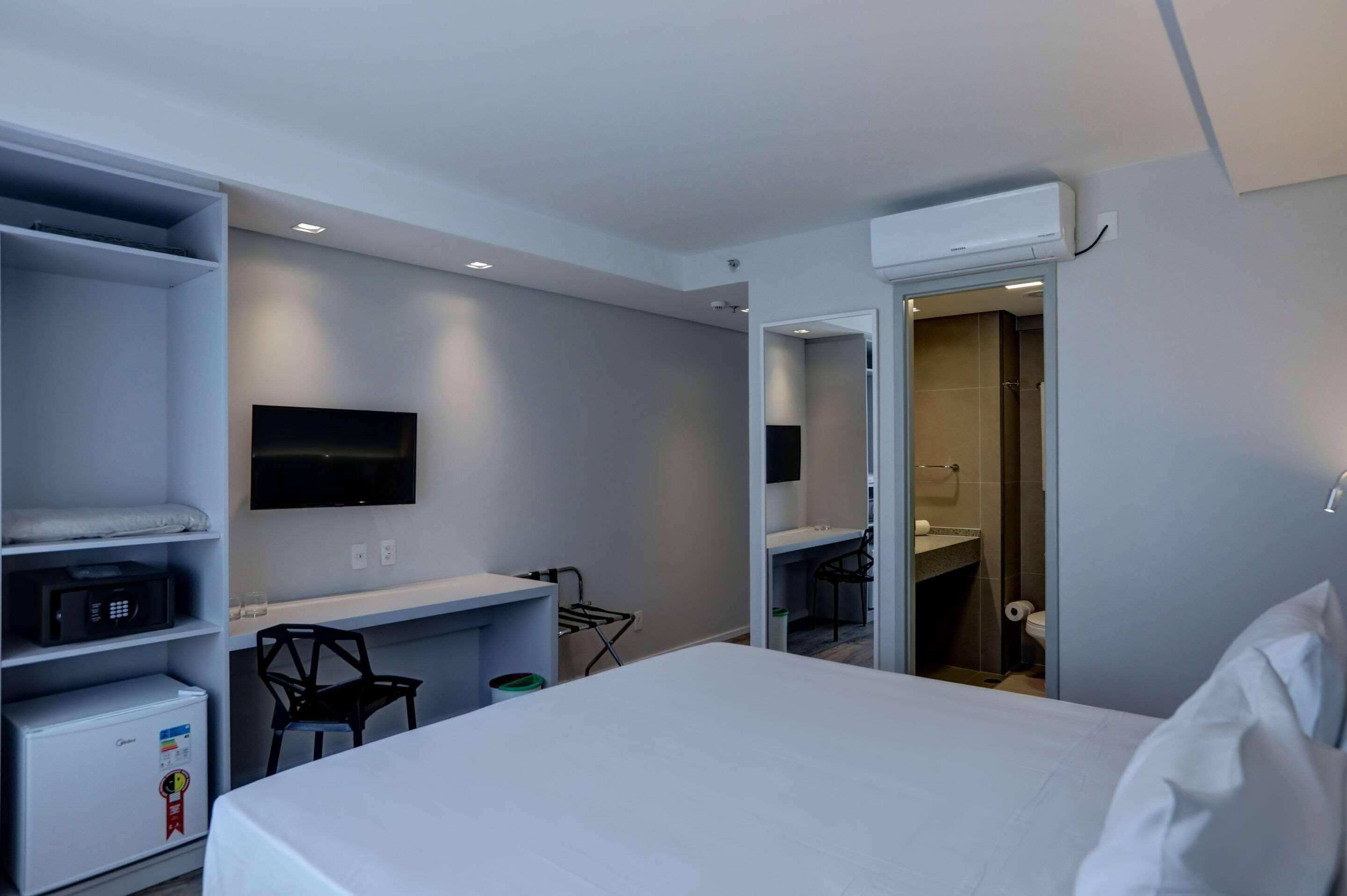 Una habitación de hotel moderna con una cama, un escritorio, una silla, un televisor de pantalla plana, un microondas, un refrigerador compacto y un baño visible a través de una puerta abierta.