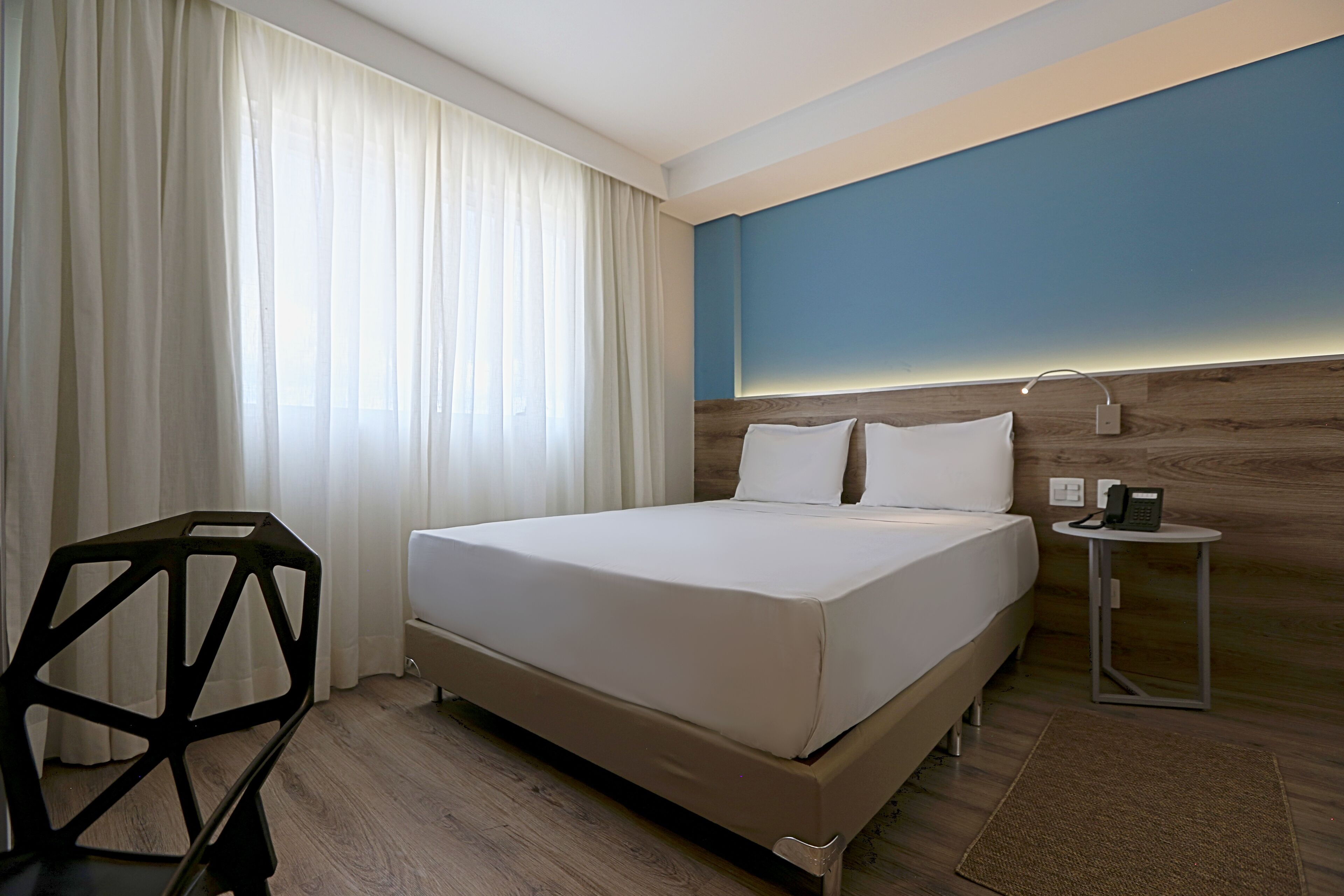 A hotel room with a bed, a desk, a chair, a phone, and a wall-mounted light.