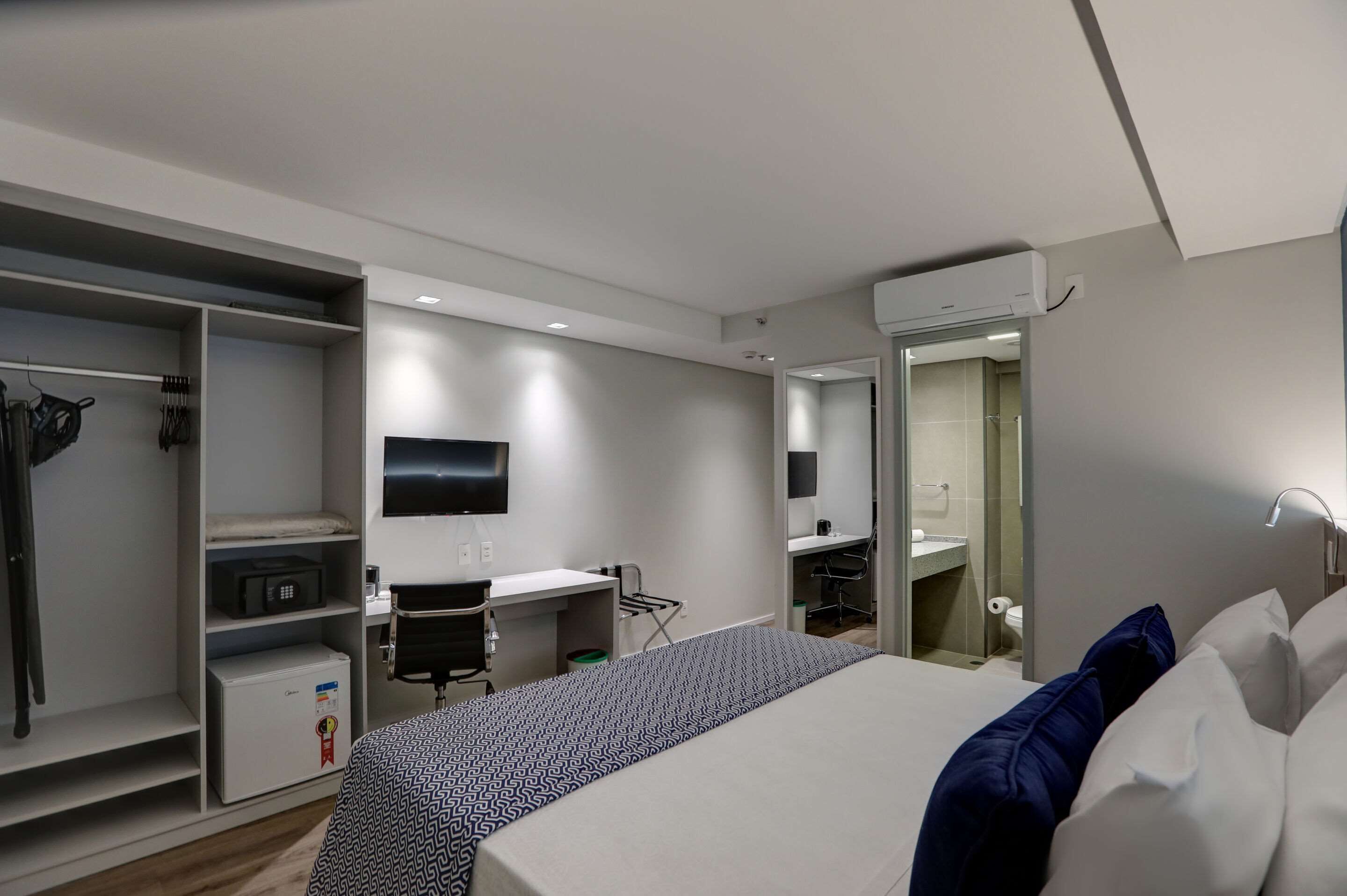 Quarto de hotel com cama, escrivaninha, guarda-roupa e banheiro.