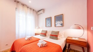 Habitación de hotel con dos camas, una mesita de noche de madera, un escritorio y una ventana con cortinas traslúcidas.