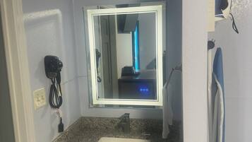 A bathroom with a granite countertop, a white sink, a mirror with a light, and a hairdryer on a shelf.
