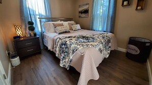 Un dormitorio con cama, mesitas de noche, lámpara, cubo de basura y ventana con cortinas.