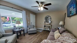 Una sala de estar con sofá, sillón y ventilador de techo. Se ve una ventana con cortinas, una puerta y un televisor montado en la pared.