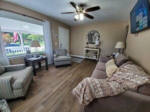 A living room with a sofa, armchair, and a ceiling fan. There is a window with curtains, a door, and a TV mounted on the wall.