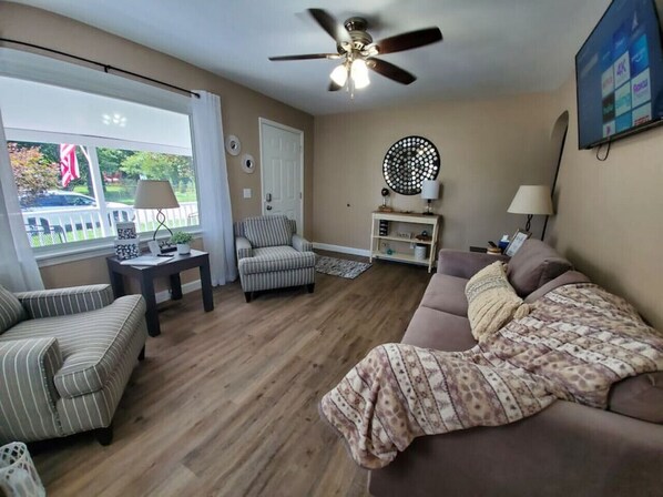 A living room with a sofa, armchair, and a ceiling fan. There is a window with curtains, a door, and a TV mounted on the wall.