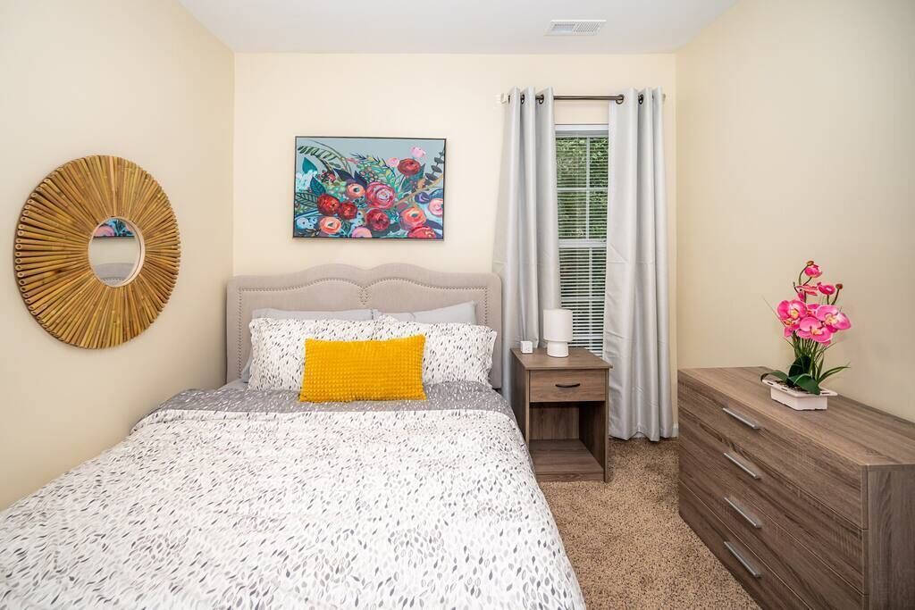 Un dormitorio con cama, mesita de noche, cómoda, espejo y un cuadro.