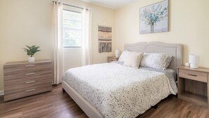 Een slaapkamer met een bed, nachtkastjes, een kaptafel, een raam met gordijnen en een aan de muur hangend schilderij.