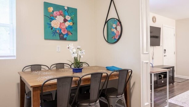 Een eetruimte met een houten tafel en metalen stoelen, een schilderij van bloemen aan de muur en een kleine potplant op tafel.