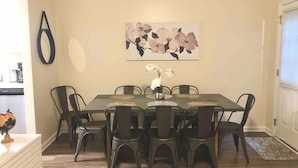 Un comedor con una mesa rectangular rodeada por seis sillas, un florero con flores sobre la mesa, y un cuadro grande con motivos florales en la pared.