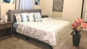 Un dormitorio con cama, mesitas de noche, lámpara, un florero con flores y decoración en la pared.