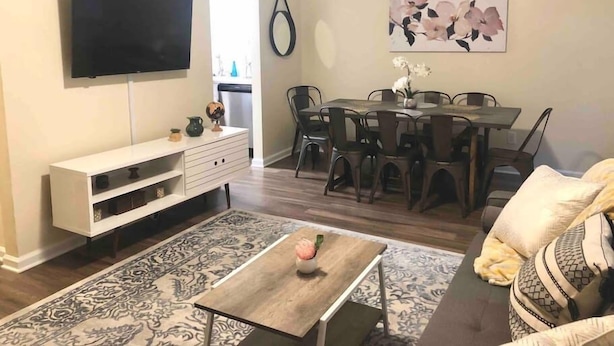Una sala de estar con un televisor de pantalla plana, un mueble blanco para equipo audiovisual, una mesa de comedor con sillas y una alfombra estampada.