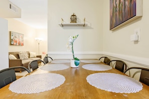 Un coin repas avec une table en bois, des sets de table blancs et un vase rempli de fleurs. On y trouve également un canapé et un tableau accroché au mur.