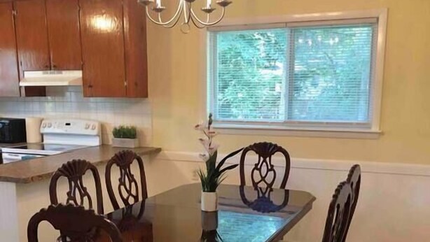 A dining area with a wooden table and chairs, a chandelier, a window with blinds, and wooden cabinets.