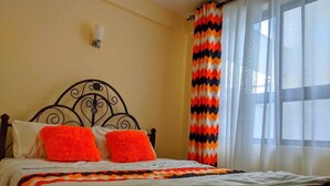 Un letto con struttura in metallo nero, biancheria da letto bianca e due cuscini arancioni. Si vede una finestra con tende bianche e una tenda colorata a zigzag sulla destra.