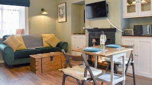 Un salon chaleureux avec un canapé vert, une commode en bois et une table à manger avec des chaises. On aperçoit une cheminée et des meubles de cuisine au fond.