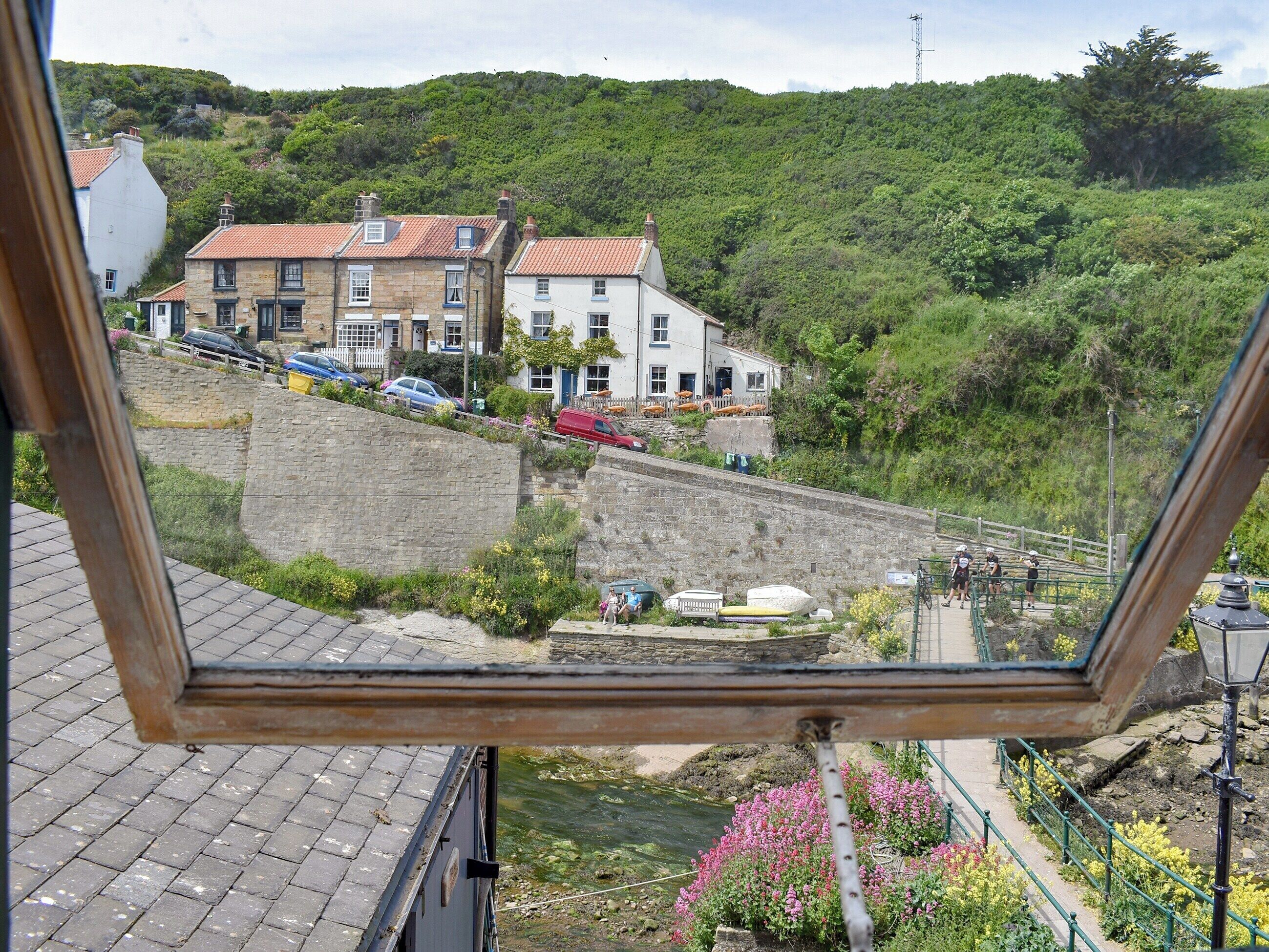 Une vue depuis une fenêtre donnant sur une colline, avec des bâtiments, un mur de pierre et un sentier où l’on voit des personnes se promener.