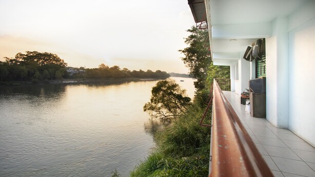 Une vue sur la rivière, avec un banc donnant sur l’eau, un bâtiment doté d’un balcon et des arbres en arrière-plan.