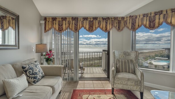 Een woonkamer met een bank, fauteuil en uitzicht op het strand door grote ramen.