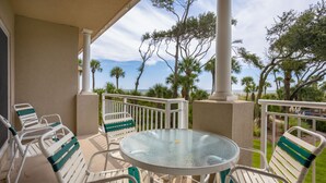 A covered outdoor seating area with a glass table and chairs, surrounded by palm trees and a view of the ocean. - Family Villa | Ocean/Pool Views, Balcony, Bikes, Close to Shops/Dining (Hilton Head Island)