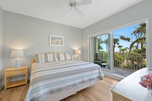A bedroom with a bed, two bedside tables with lamps, a ceiling fan, a painting, and a view of palm trees through a sliding glass door. - Family Villa | Ocean/Pool Views, Balcony, Bikes, Close to Shops/Dining (Hilton Head Island)