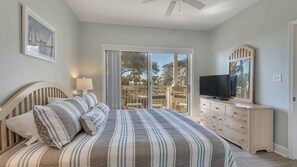 Ein Schlafzimmer mit Bett, Kommode, Fernseher und einem Balkon mit Blick auf Gebäude und Bäume.