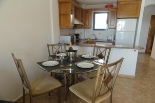 Un comedor con una mesa redonda de cristal dispuesta para seis personas, rodeada de sillas de madera. Una botella de champán está colocada sobre la mesa. Se observa una cocina al fondo con armarios de madera y un refrigerador blanco.