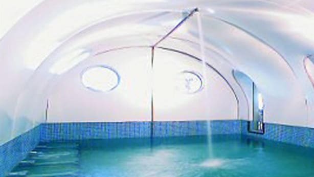 Une piscine intérieure dotée d’un sol et de murs recouverts de carreaux bleus, d’un plafond en forme de dôme et de deux fenêtres circulaires.