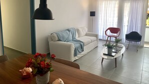 Uma sala de estar com um sofá branco, uma cadeira vermelha, uma mesa de centro com uma planta e uma mesa de jantar de madeira com um vaso de flores.