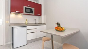 Une cuisine compacte avec des meubles blancs, un micro-ondes rouge, un petit réfrigérateur et un panier de fruits posé sur le plan de travail.