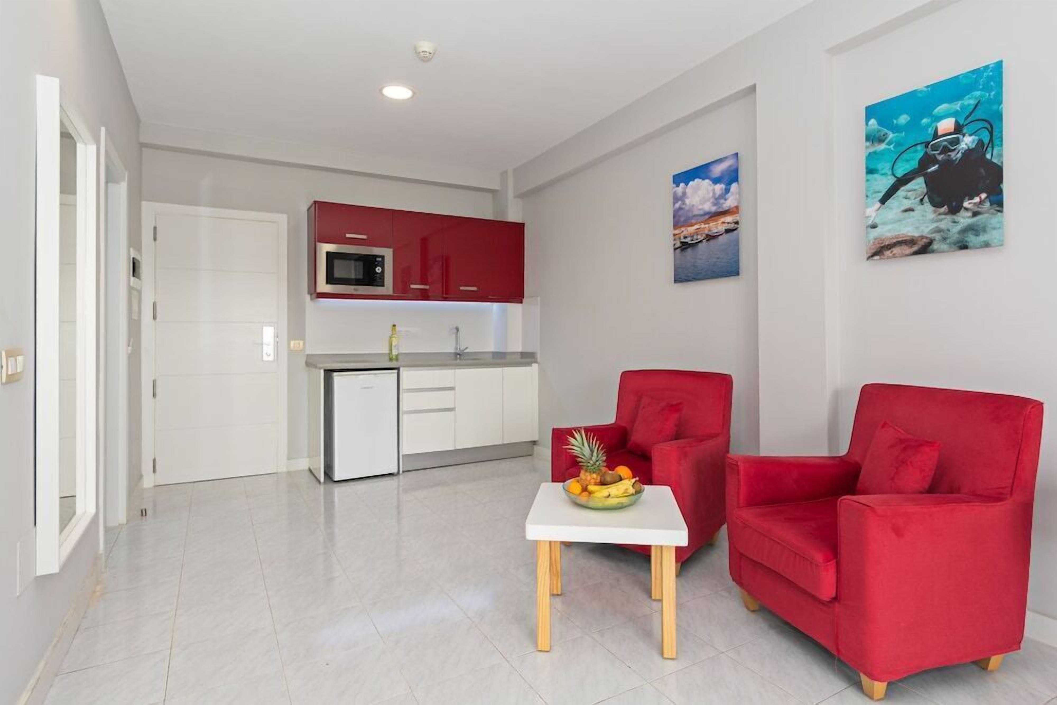 Una sala de estar moderna con un sofá rojo, una mesita blanca con fruta y una cocina al fondo.