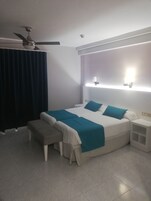 Un dormitorio moderno con una cama grande, mesitas de noche, un banco, un ventilador de techo y una lámpara fijada a la pared.