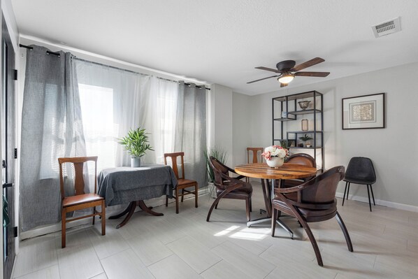 Un coin repas comprenant une table ronde en bois, quatre chaises, un ventilateur de plafond et une bibliothèque agrémentée d’objets décoratifs.