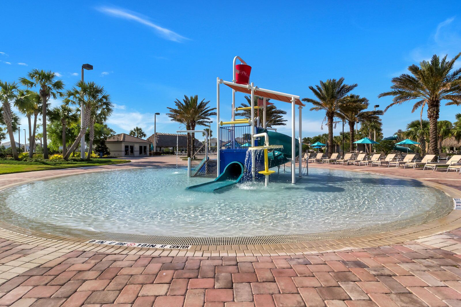 A water play area with a slide and a red bucket, surrounded by palm trees and lounge chairs.