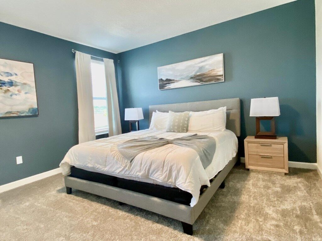A bedroom with a bed, two bedside tables with lamps, a window with curtains, and two wall-mounted paintings.