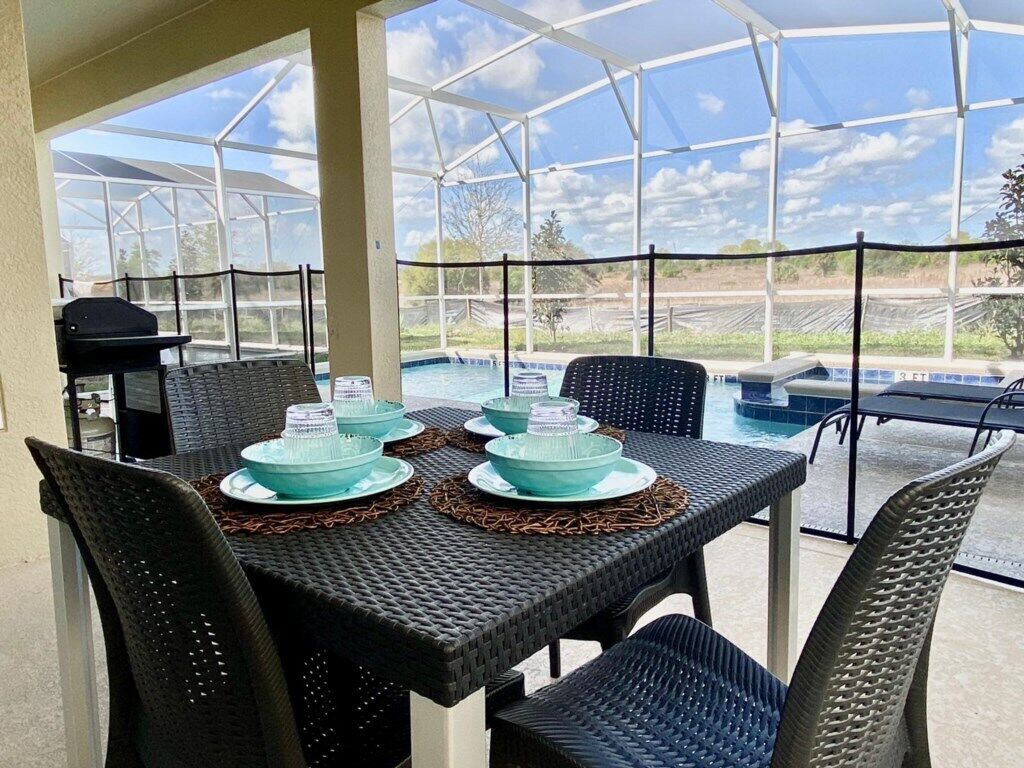 Une terrasse extérieure avec une table dressée pour quatre personnes, entourée de chaises en osier, donnant sur une piscine et un jardin.