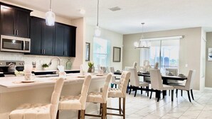 A modern kitchen with dark cabinets, a central island with chairs, and a dining area with a large window. - Luxury 6 Bedroom 5 bath pool Home 202 (Four Corners)